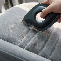 A hand holds a black pet grooming brush over a gray upholstered car seat, showing loose fur being removed.