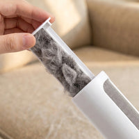 A close-up of a hand holding a transparent cylindrical filter or container filled with gray pet fur, shown against a beige sofa background.